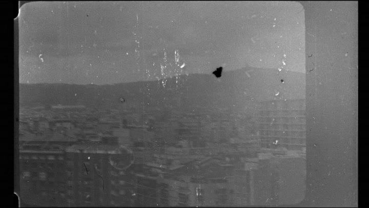 Un fotogramma in 16 mm, in bianco e nero: la citt&agrave; dall&rsquo;alto e le colline, con la polvere e i bordi della pellicola.